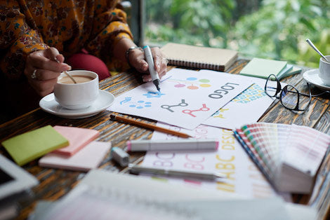 A person creating designs for a logo while sitting at a desk and stirring their coffee