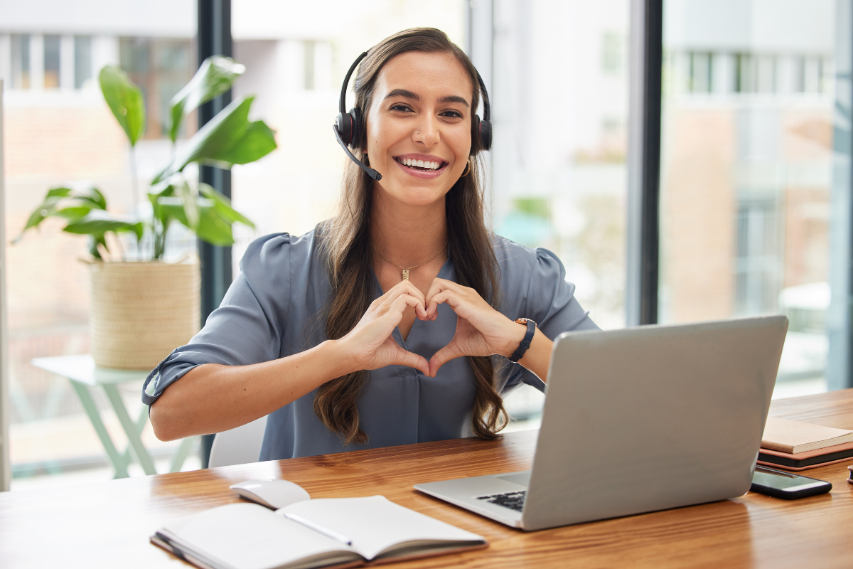 A smiling woman sitting at her laptop holding up her hands in the shape of a heart